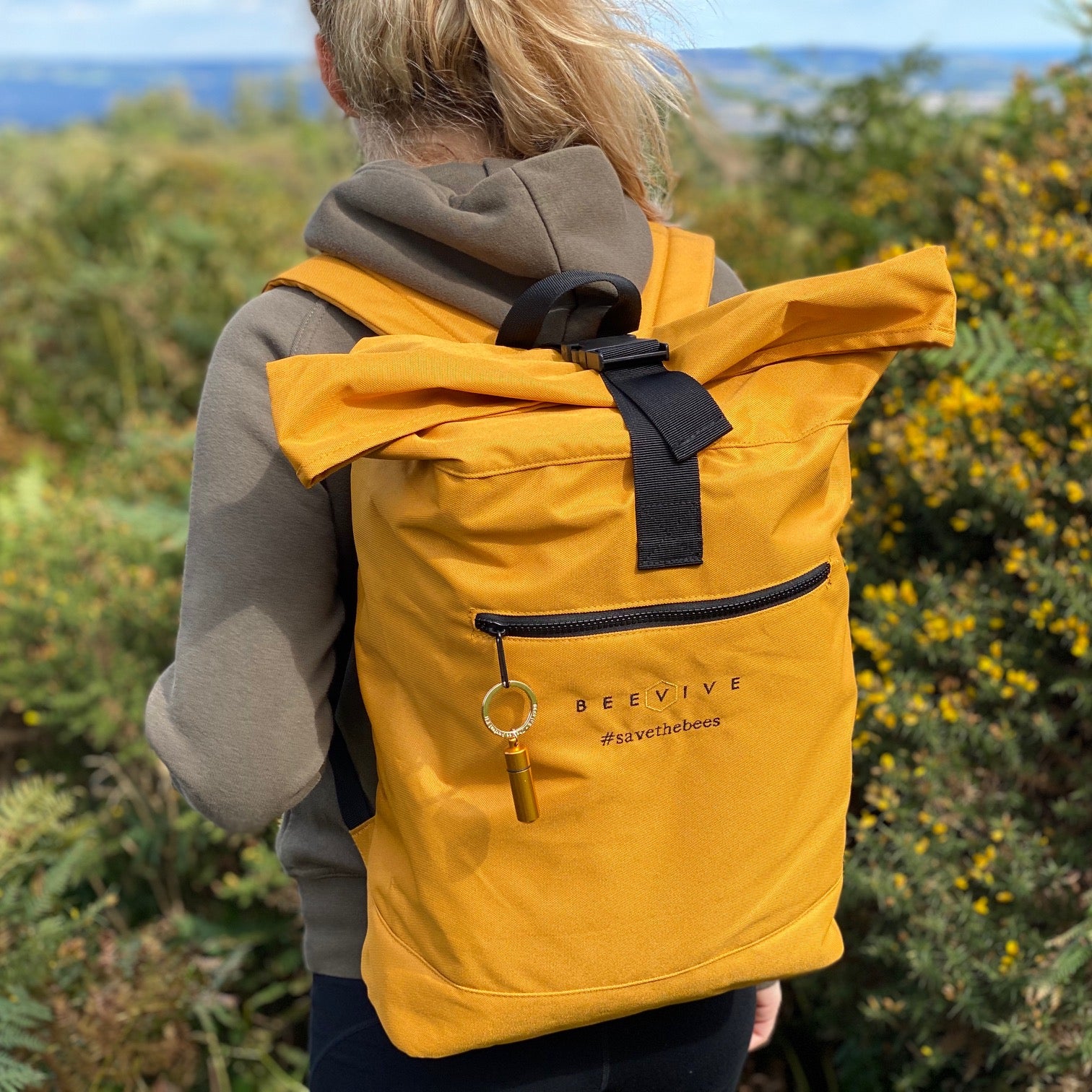 young female walking amongst flowering gorse wearing a mustard colour Beevive adventure recycled roll-top backpack. gold bee revival kit attached to the zipper - backpack shows hashtag 'savethebees' and the beevive logo stitching