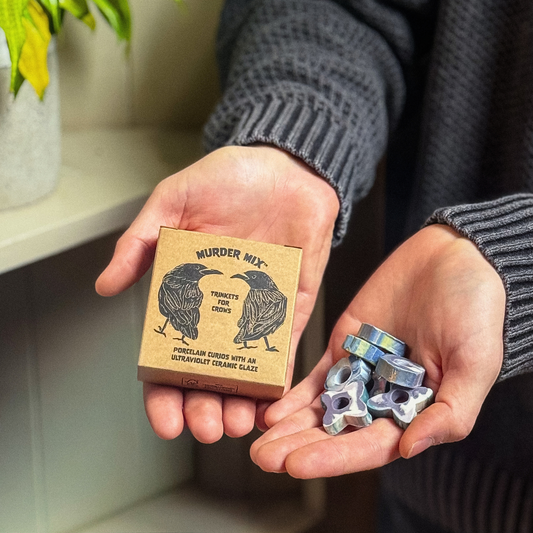 A person holds a small printed box labeled 'Murder Mix' next to their hands, which hold several metallic star-shaped ceramic grinding carbides. The background shows a softly blurred indoor setting with a shelf and plant.