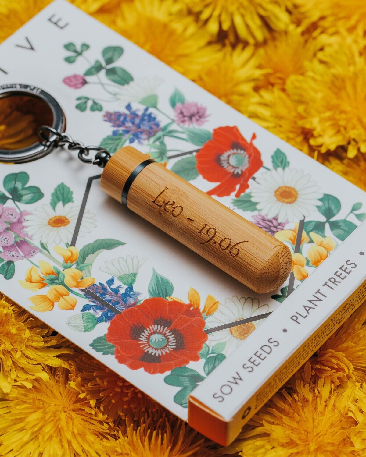 anthracite grey bamboo bee revival keyring still in its packaging resting on bed of yellow flower heads