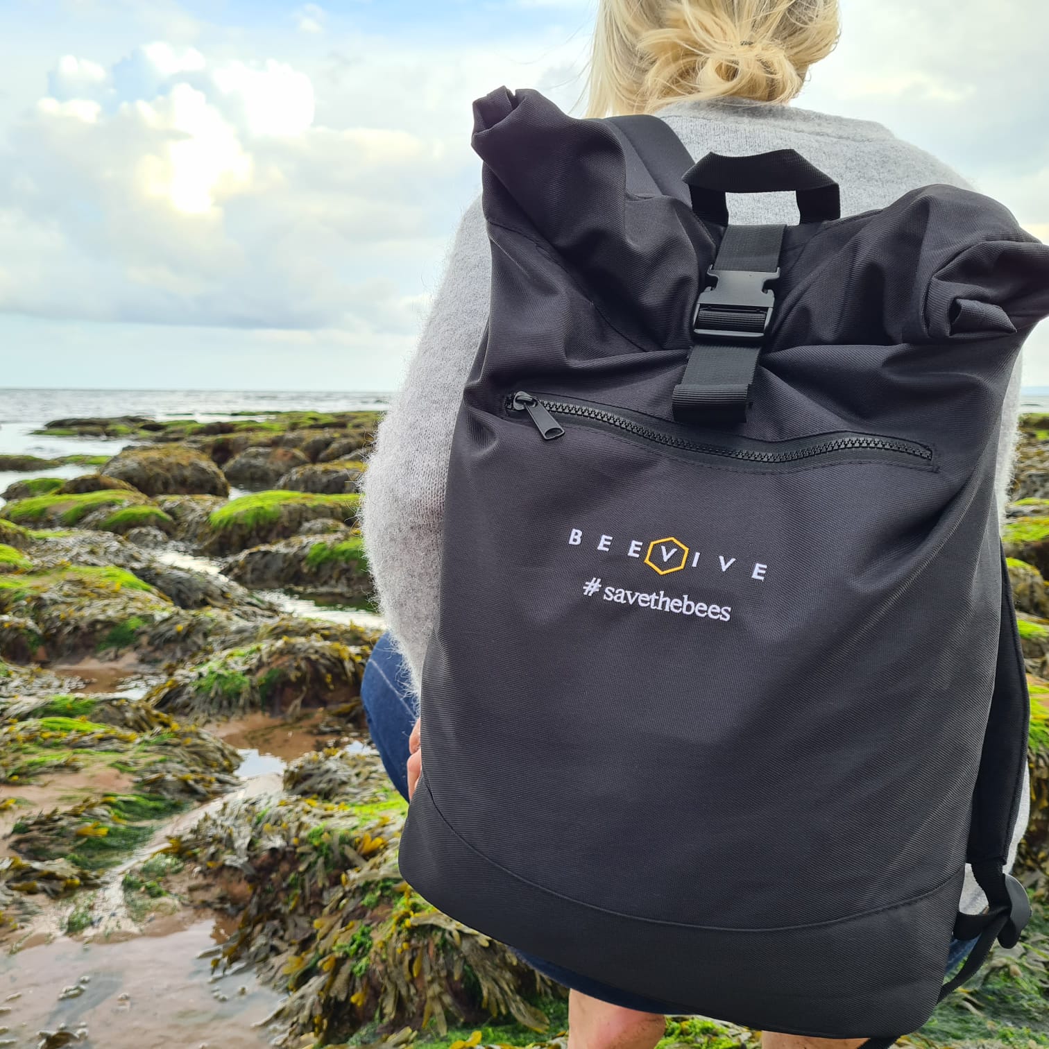 young female walking across rocks covered in seaweed wearing the black coloured Beevive adventure recycled roll-top backpack. - backpack shows hashtag 'savethebees' and the beevive logo stitching